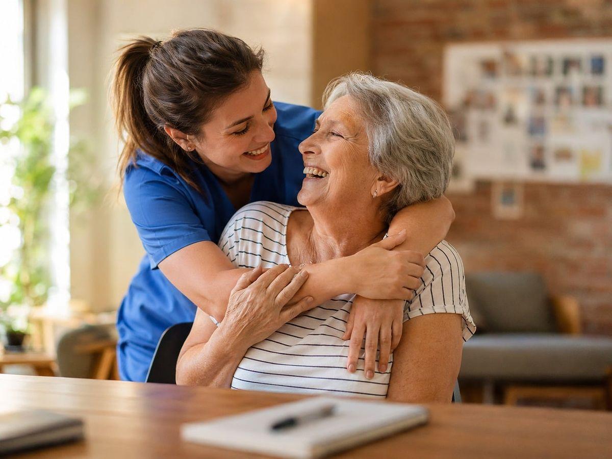 Caregiver in blue scrubs hugs an elderly woman, both smiling in a cozy living room setting.